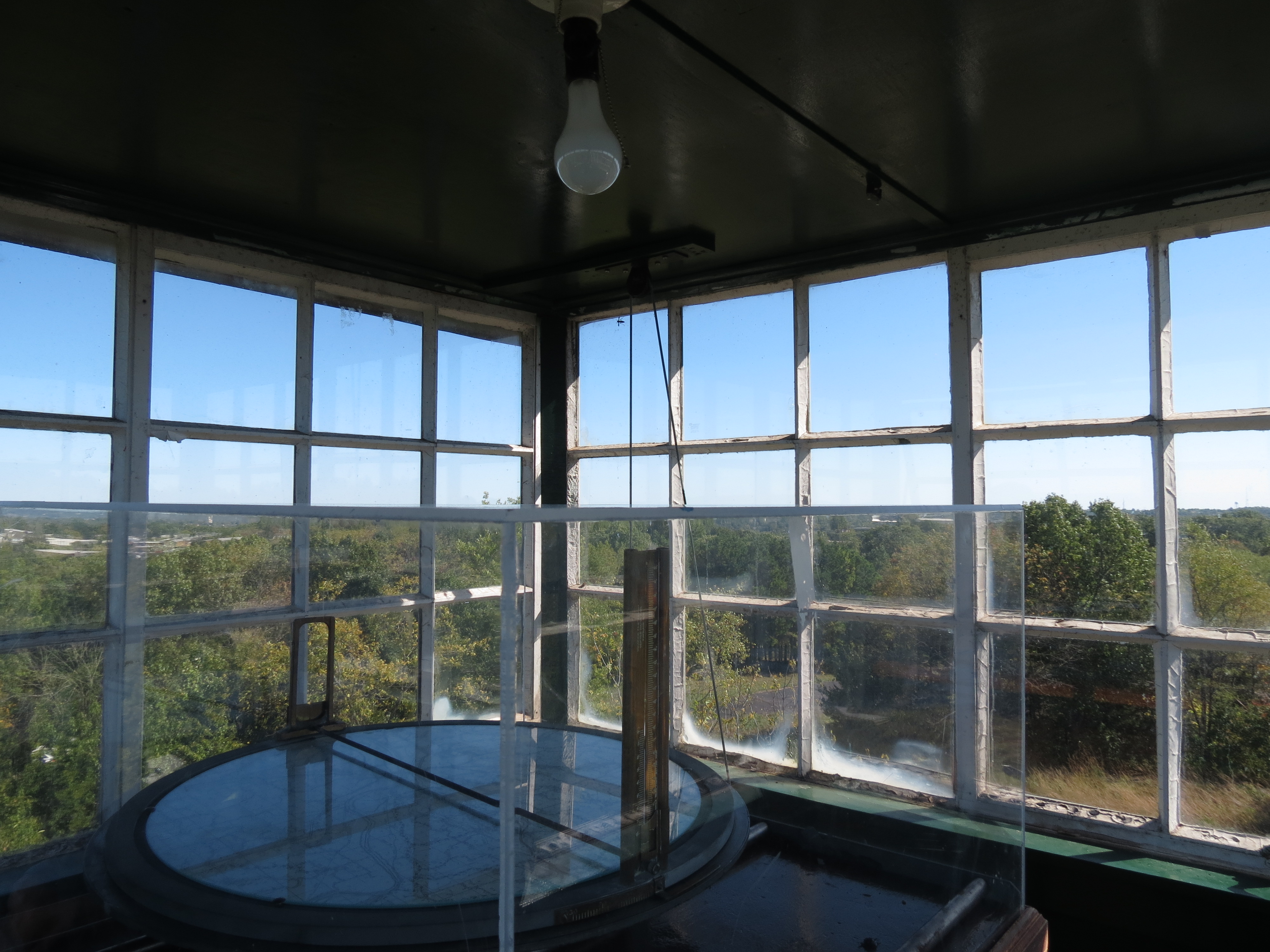 The Osborne Fire Finder sitting in a glass case inside of the fire tower with the trees in the background.