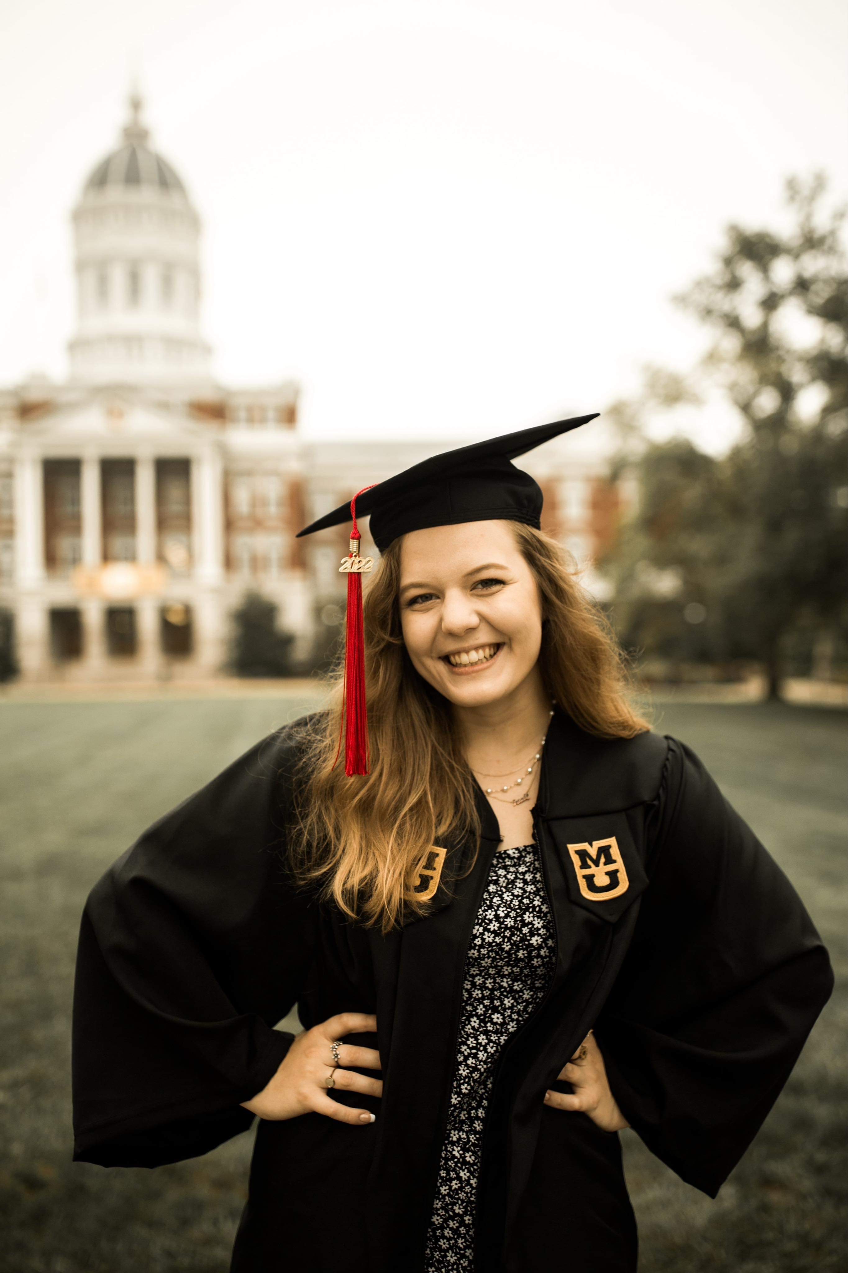 Katrina Troy, a blonde young woman, stands in front of Jesse Hall on the Francis Quad in her graduation gown and cap. She has red tassle with a 2022 charm.