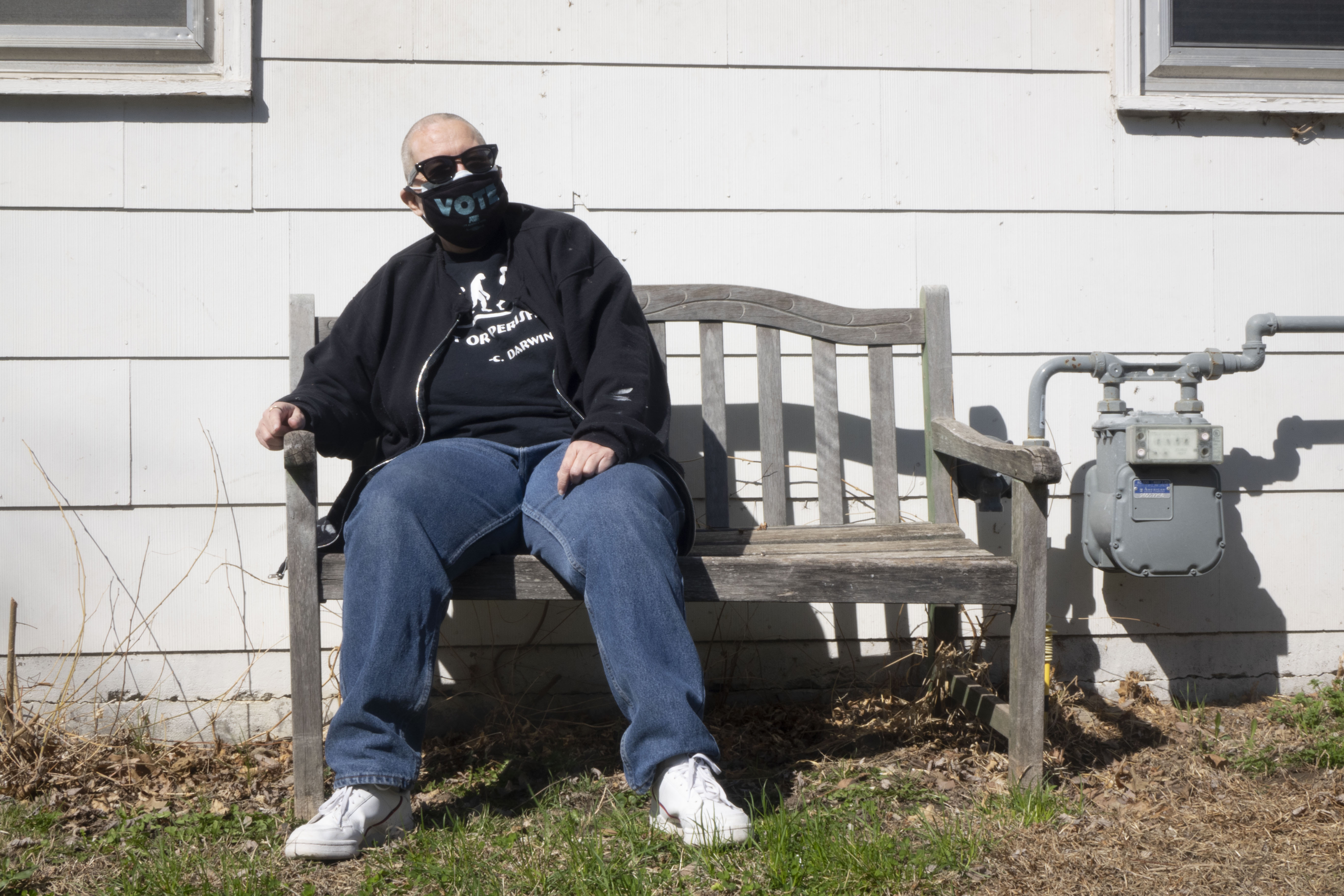 Columbia Activist Rene Powell is sitting on a small wooden bench outside her home. She is wearing black sunglasses, a black mask that reads, vote, and a black shirt that reads, adapt or perish, a quote by Charles Darwin.